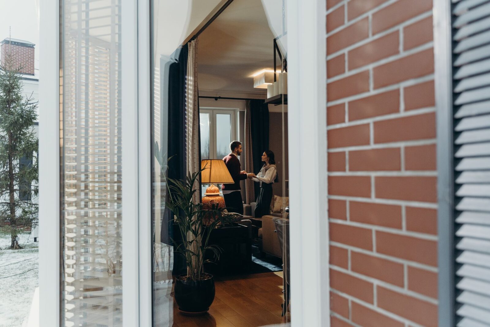 A couple stands in a stylish room seen through a window, holding hands.